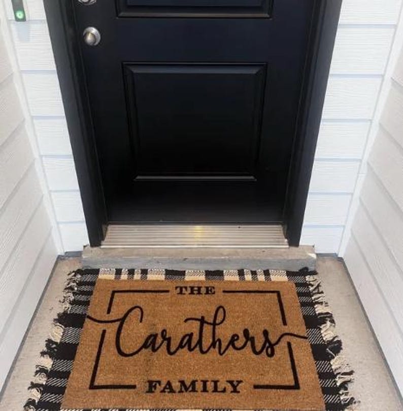 May include: A rectangular doormat with the text "THE Carathers FAMILY" in black script and block letters. The mat is brown with a black and white buffalo plaid border and fringe. The mat is placed in front of a black door.