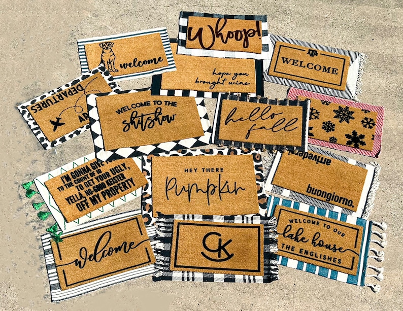 May include: A collection of welcome mats with various designs and sayings. Some mats feature a black and white checkered pattern, while others have a more rustic look. The mats include sayings such as "Welcome to the Shitshow", "Hey There Pumpkin", "Welcome to Our Lake House", and "Buongiorno".