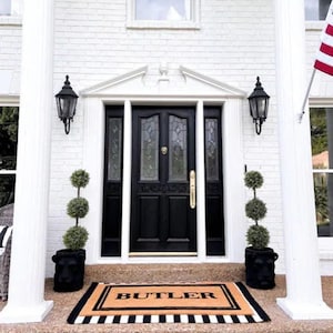 May include: A black front door with a glass panel and a gold door handle. The door is flanked by two black lanterns and two potted topiary trees. A welcome mat with the word "BUTLER" in black lettering is on the front porch.