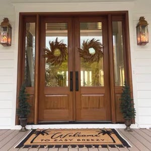 May include: Double wooden doors with glass panels and black door handles. The doors are decorated with two dried grass wreaths. A welcome mat with the text "Welcome to Easy Breezy" is placed in front of the doors.