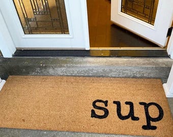 May include: A brown coir doormat with the word "sup" in black lettering.