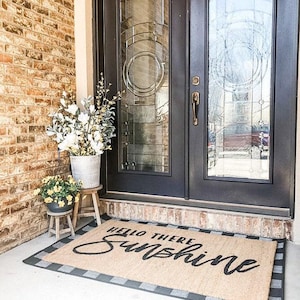 Puede incluir: Una alfombra de bienvenida con un borde a cuadros blanco y negro y el texto "Hello There Sunshine" en letra cursiva negra. La alfombra se coloca delante de una doble puerta negra con paneles de vidrio.