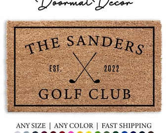 May include: A brown coir doormat with black text and a black outline. The text reads "The Sanders Golf Club Est. 2022" with two crossed golf clubs.