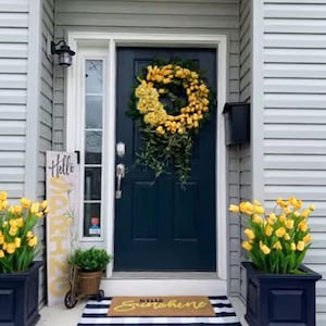 Puede incluir: Una puerta delantera azul con una corona de flores amarillas, un letrero de bienvenida que dice "Hello Sunshine" y dos macetas con flores amarillas a cada lado de la puerta.