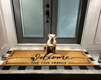 May include: A tan doormat with the word "Welcome" in cursive and "THE COX FAMILY" in a block font. A brown and white bulldog sits on the mat. The mat is bordered by a black and white checkered pattern.