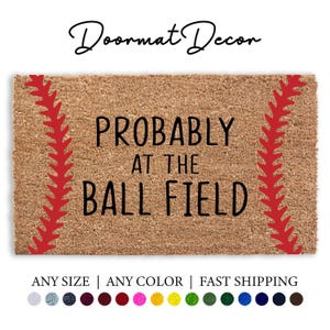 May include: A brown coir doormat with red stitching around the edges, resembling a baseball. The text "PROBABLY AT THE BALL FIELD" is printed in black on the mat.