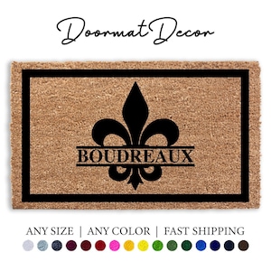 Op de afbeelding: Een rechthoekige deurmat met een zwarte rand en een fleur-de-lis-ontwerp. Het woord "BOUDREAUX" is gecentreerd onder de fleur-de-lis. De tekst "Doormat Decor" staat in cursief bovenaan. De deurmat is bruin.