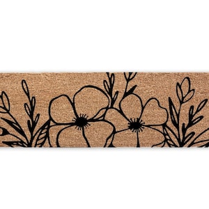 Puede incluir: Felpudo rectangular de fibra de coco marrón natural. Presenta un diseño floral negro con dos flores grandes y detalles de hojas. El diseño está centrado, creando una entrada acogedora.