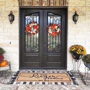 May include: A welcome mat with black and white checkered trim and the text "The Rieger Family Est. 2018" in a cursive font. The mat is placed in front of a double black door with decorative ironwork and two fall wreaths.