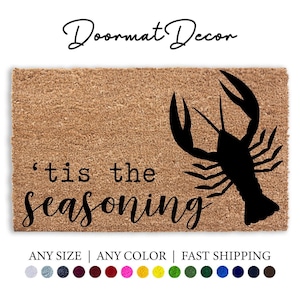 May include: A brown coir doormat with the text "'tis the seasoning" in black cursive font. A black silhouette of a lobster is on the right side of the mat.