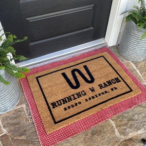 May include: A rectangular doormat with the text "RUNNING W RANCH ZOLFO SPRINGS, FL" in black on a tan background. The mat is framed with a black border and a red and white polka dot border. Two galvanized metal planters with ferns flank the door.