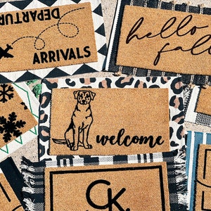 May include: A collection of eight doormats with various designs, including "Departures Arrivals", "Hello Fall", "Welcome", "Lake", and a dog silhouette. The doormats are made of coir and have a natural brown color.