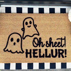 Könnte beinhalten: Eine braune Kokosfaser-Fußmatte mit schwarz-weiß gestreiftem Rand. Die Fußmatte hat zwei schwarze Geisterumrisse und den Text "Oh sheet! HELLUR!" in Schwarz.