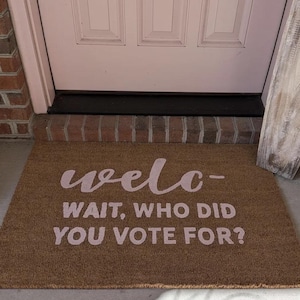 May include: A rectangular doormat made of natural fibers, featuring the word "welc-" above the phrase "WAIT, WHO DID YOU VOTE FOR?" in a light pink color. The mat is placed in front of a white door.