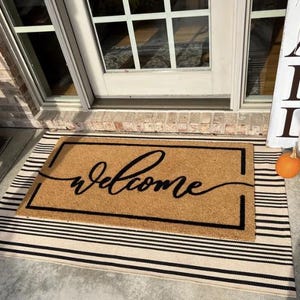 Peut inclure: Un paillasson en fibre de coco brun avec une bordure noire et le mot "welcome" écrit en cursive. Le paillasson est sur un tapis rayé noir et blanc.