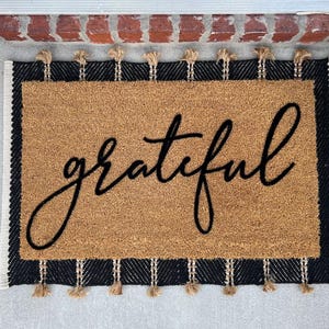 May include: A brown coir doormat with black and white trim and tassels. The word "grateful" is written in black cursive lettering on the mat.