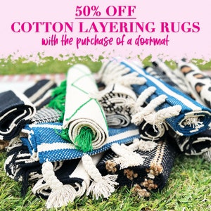 May include: A stack of rolled cotton rugs in various patterns and colors, including black, white, blue, and green. The rugs have tassels and are arranged on a bed of green grass. The text "50% OFF COTTON LAYERING RUGS with the purchase of a doormat" is displayed above the rugs.