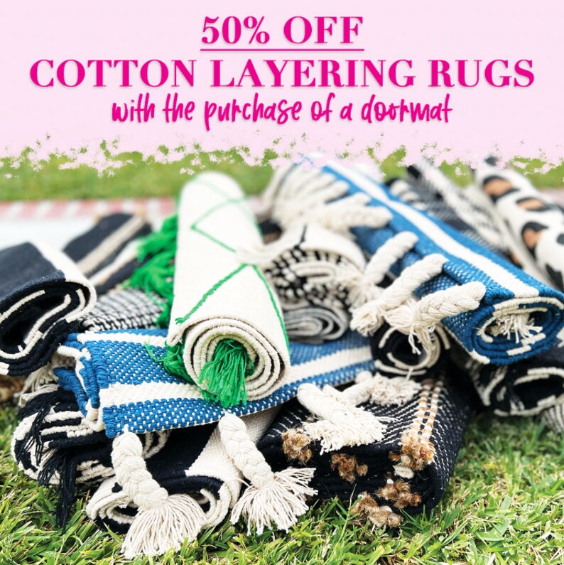 May include: A stack of rolled cotton rugs in various patterns and colors, including black, white, blue, and green. The rugs have tassels and are arranged on a bed of green grass. The text "50% OFF COTTON LAYERING RUGS with the purchase of a doormat" is displayed above the rugs.