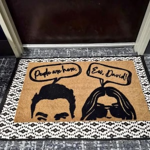 May include: A rectangular doormat with a brown center and a black and white patterned border. The mat features black silhouettes of two people with speech bubbles that read "People are here." and "Ew, David!"