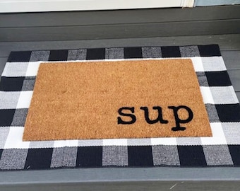 May include: A rectangular doormat with the word "sup" in black letters. The doormat is brown and sits on top of a black and white checkered rug.