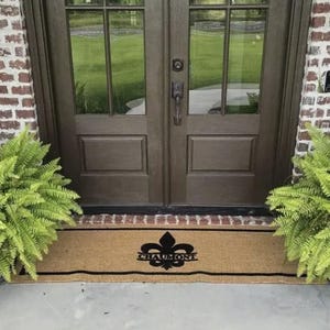 Op de afbeelding: Een rechthoekige kokosmat met een zwart fleur-de-lis ontwerp en de naam "CHAUMONT". De mat ligt voor een bruine dubbele deur met glazen panelen, geflankeerd door groene varens en metselwerk.