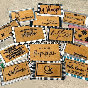 May include: A collection of welcome mats with various designs and sayings. Some mats feature a black and white checkered pattern, while others have a more rustic look. The mats include sayings such as "Welcome to the Shitshow", "Hey There Pumpkin", "Welcome to Our Lake House", and "Buongiorno".