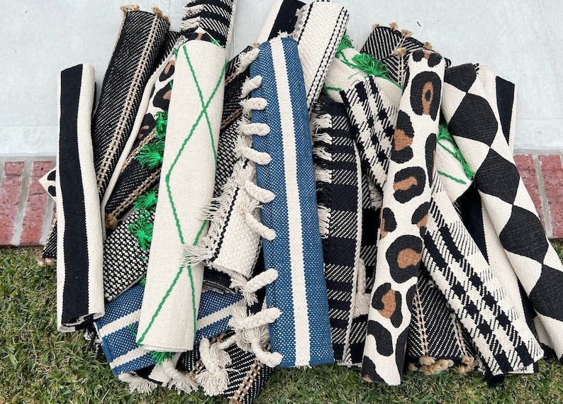 May include: A collection of rolled-up rugs with various patterns, including black and white stripes, black and white checkered, leopard print, and a geometric design with green accents. The rugs are made of natural fibers and have fringe edges.