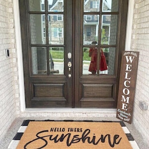Puede incluir: Una puerta de madera marrón con un letrero de bienvenida que dice "Welcome Dambach Home". Una alfombra de bienvenida con el texto "Hello There Sunshine" está delante de la puerta.