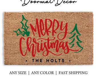 Könnte beinhalten: Eine braune Kokosmatte mit dem Text "Merry Christmas" in roter Kursivschrift und grünen Stechpalmen- und Kiefernbaum-Akzenten. Der Text "The Holts" ist in Schwarz unter dem Haupttext geschrieben.