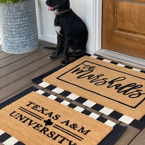 May include: Two doormats with black and white checkered borders. The top doormat is brown with the text "the marshalls" in black cursive. The bottom doormat is brown with the text "TEXAS A&M UNIVERSITY" in black. A black dog is sitting on the porch in front of the doormats.