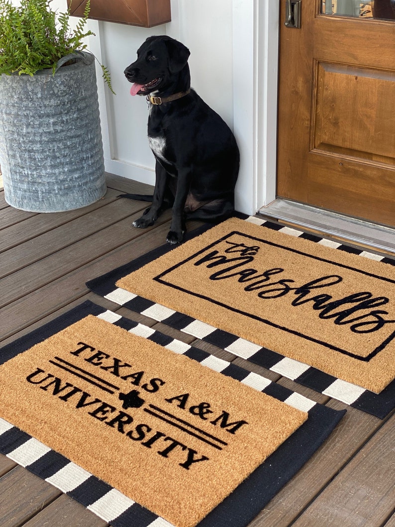 May include: Two doormats with black and white checkered borders. The top doormat is brown with the text "the marshalls" in black cursive. The bottom doormat is brown with the text "TEXAS A&M UNIVERSITY" in black. A black dog is sitting on the porch in front of the doormats.