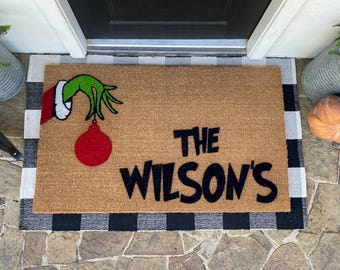 May include: A brown coir doormat with a black and white chequered border. The doormat features a green Grinch hand holding a red bauble and the text "The Wilson's".