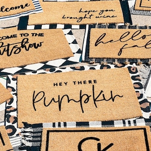 May include: A collection of doormats with various welcome messages, including "Welcome to the shitshow", "Hope you brought wine", "Hello Fall", "Hey there Pumpkin", and "Ugly, Sister Property".