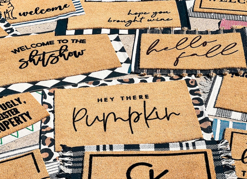 May include: A collection of doormats with various welcome messages, including "Welcome to the shitshow", "Hope you brought wine", "Hello Fall", "Hey there Pumpkin", and "Ugly, Sister Property".