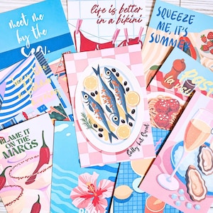 May include: A collection of colorful greeting cards with summer and food-themed illustrations. Cards feature phrases like "Meet me by the sea," "Life is better in a bikini," and "Squeeze me, it's summer." Other cards depict food like fish, pasta, and margaritas.