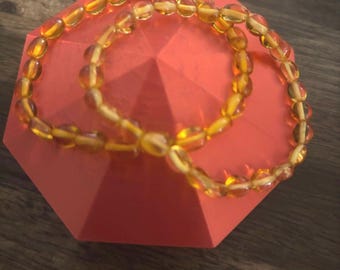 May include: Two amber beaded bracelets with oval-shaped beads, displayed on a red octagonal stand.