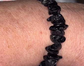 May include: A black gemstone bracelet composed of irregularly shaped, polished stones. The bracelet is strung together and worn on a wrist, showcasing the dark, reflective surfaces of the stones. The bracelet is a piece of jewelry.