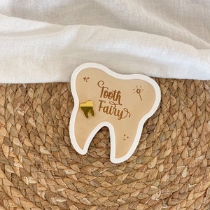 Puede incluir: Placa de madera con forma de diente con las palabras "Tooth Fairy" en fuente cursiva. La placa tiene un diente dorado y está delineada en blanco. También se incluyen estrellas decorativas y una varita.