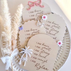 May include: Wooden affirmation signs with the phrases "I AM brave, I AM smart, I AM strong, I AM loved, I AM enough" and the name "Rylan". The signs are decorated with a pink bow, white butterflies, and flowers.