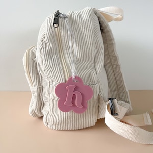 May include: A small, white corduroy backpack with a pink flower-shaped luggage tag that says "h" on it.