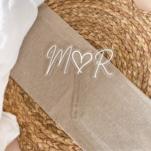 May include: White acrylic cake topper with the letters "M" and "R" with a heart in between. The letters are in a cursive font.