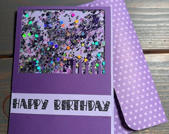 May include: Purple birthday card with a shaker window filled with glitter and confetti. The card has the words "HAPPY BIRTHDAY" in black. A matching purple envelope with white polka dots is included.