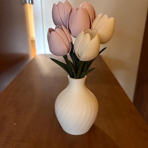 May include: A white vase with a spiral design holds a bouquet of artificial tulips. The tulips are in shades of pink and cream, with green stems and leaves. The vase sits on a wooden surface.