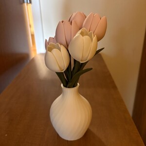 May include: A white vase with a spiral design holds a bouquet of artificial tulips. The tulips are a mix of light pink and cream colors, with green stems. The vase sits on a wooden surface.