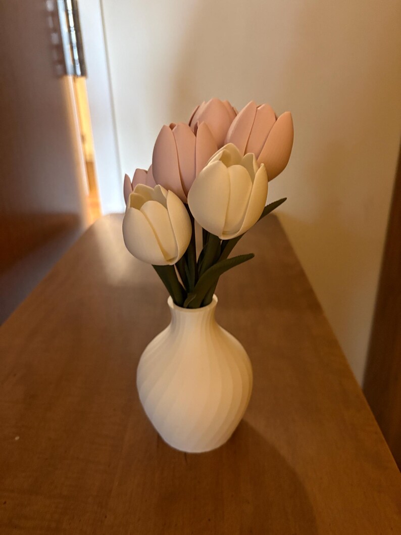 May include: A white vase with a spiral design holds a bouquet of artificial tulips. The tulips are a mix of light pink and cream colors, with green stems. The vase sits on a wooden surface.