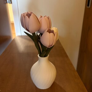 May include: A white vase with a spiral design holds a small bouquet of pink and yellow tulips with green stems. The vase sits on a wooden surface, creating a simple, elegant floral arrangement.