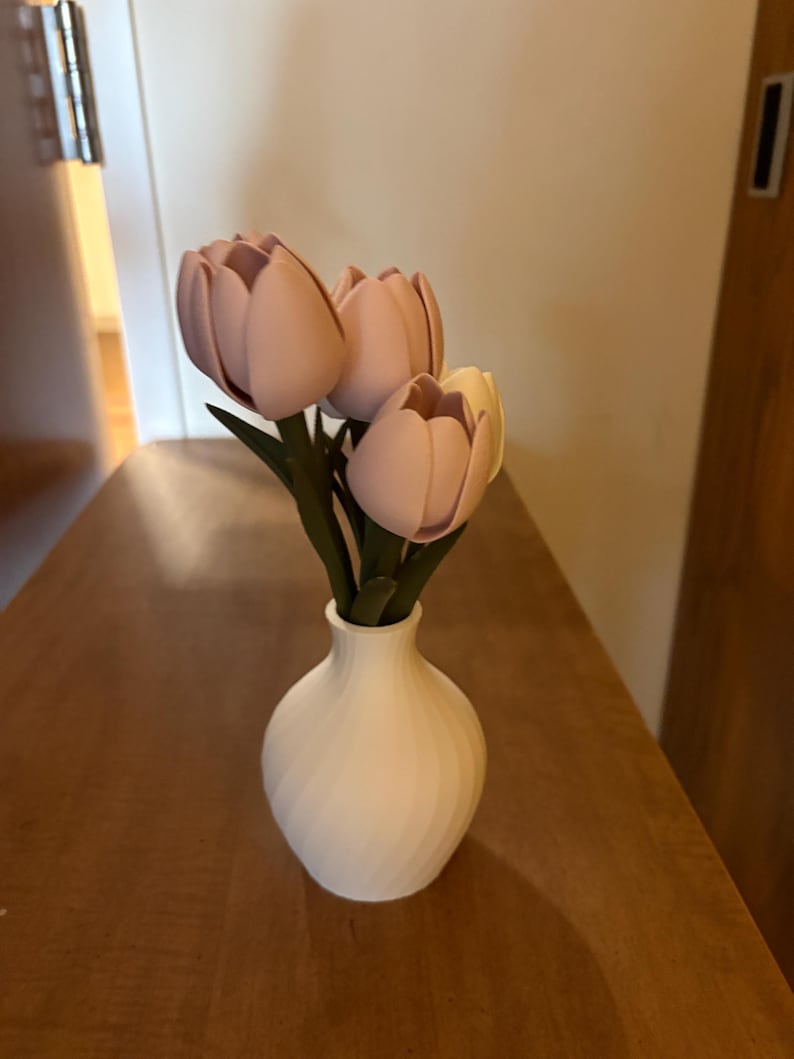 May include: A white vase with a spiral design holds a small bouquet of pink and yellow tulips with green stems. The vase sits on a wooden surface, creating a simple, elegant floral arrangement.