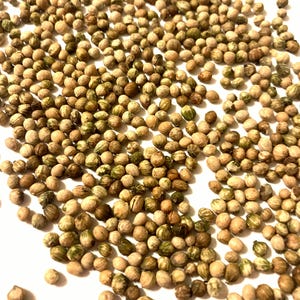May include: A close-up of a pile of coriander seeds. The seeds are light brown with green markings and are scattered on a white surface.