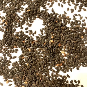 May include: Close-up of a pile of black seeds scattered on a white surface. The seeds are small, oval-shaped, and dark in color, with some showing a lighter, tan hue. The seeds are likely for culinary or gardening purposes.