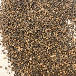 May include: Close-up of a pile of brown and black seeds. The seeds are small and round, and they are scattered on a white surface.